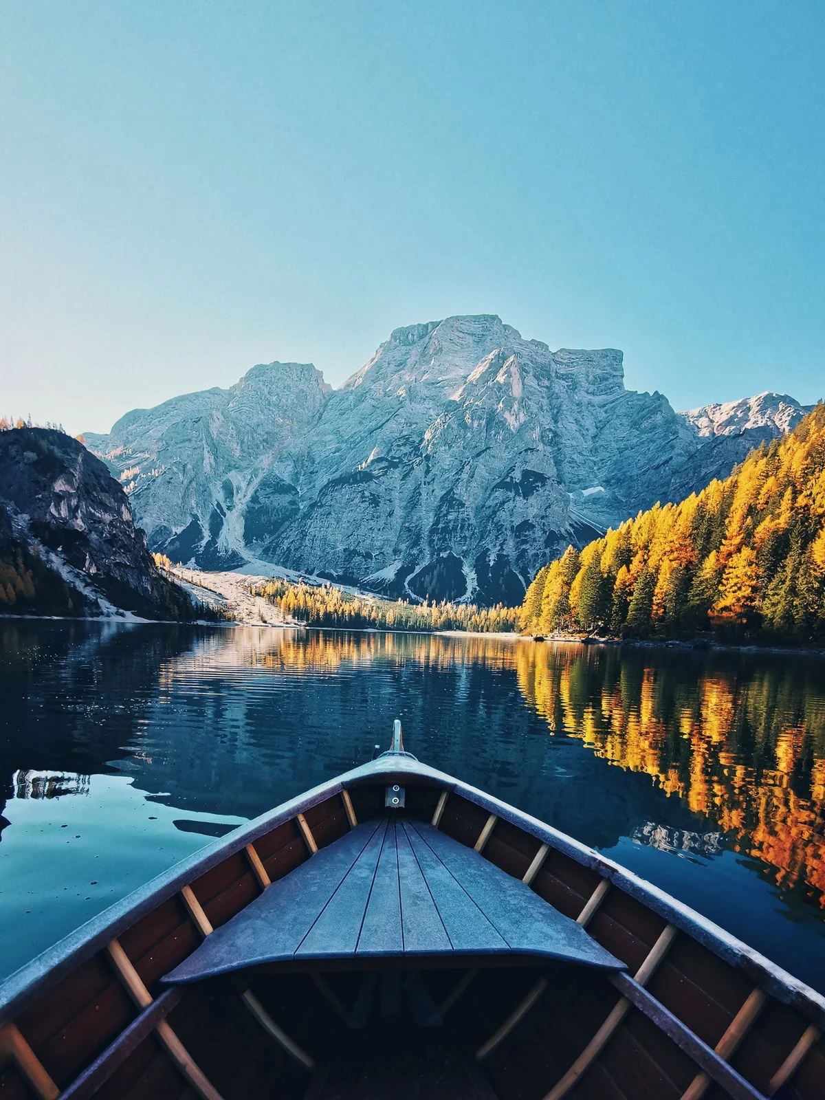 A rowboat gliding toward a towering Dolomite peak, reflected in Lake Braies beside golden autumn trees